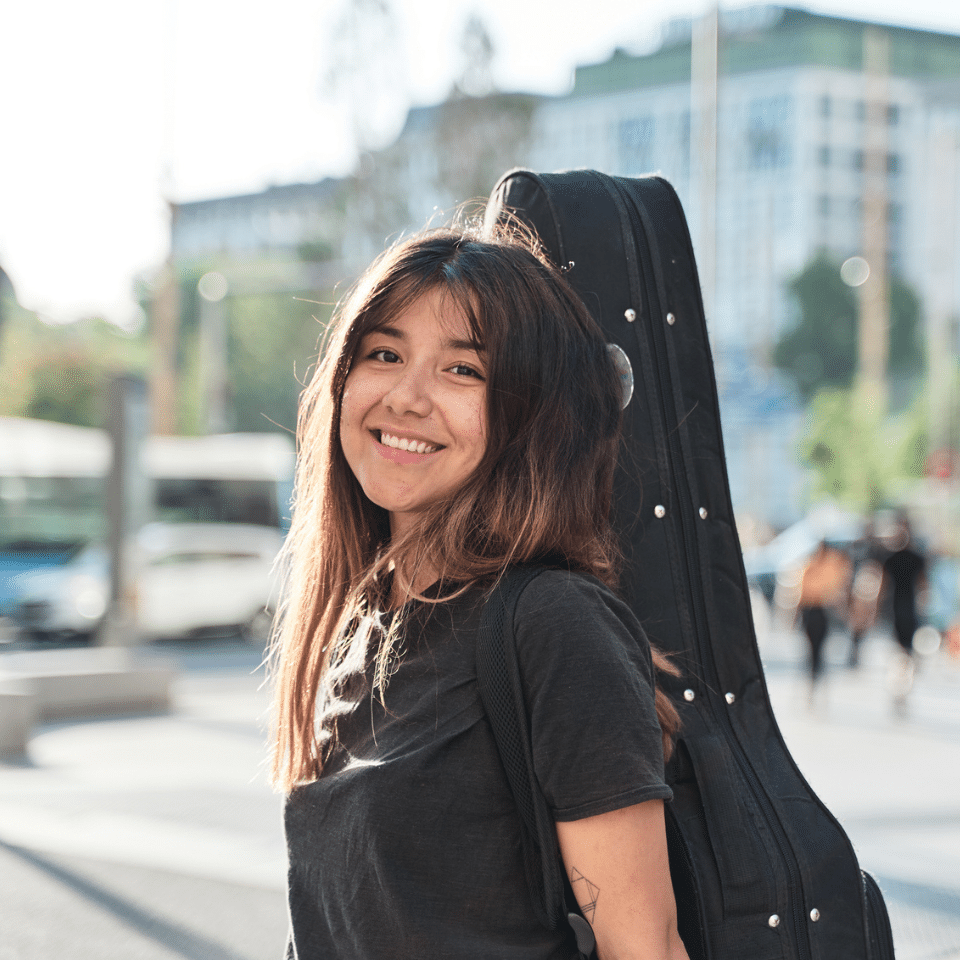 young woman smiling outside and carrying a guitar case