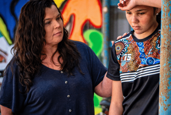 Woman comforts a teenage girl who is looking down. The girl looks serious and sad and is leaning her head against her arm.