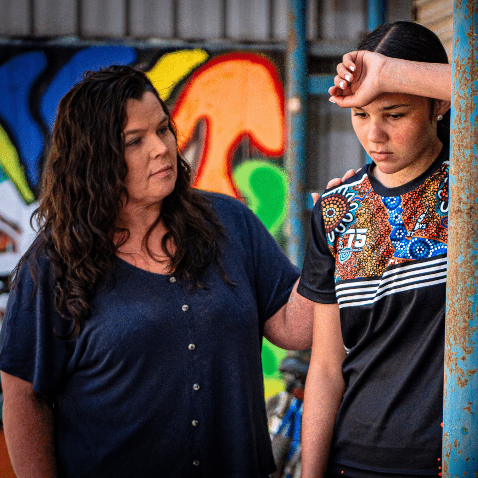 Woman comforts a teenage girl who is looking down. The girl looks serious and sad and is leaning her head against her arm.