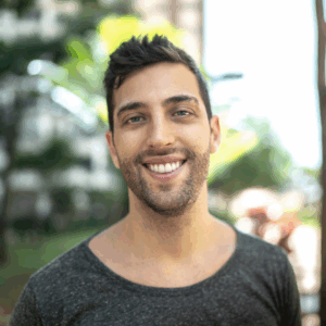 A young man smiles confidently. He is standing outside and wearing a plan grey t-shirt. The background is blurred.